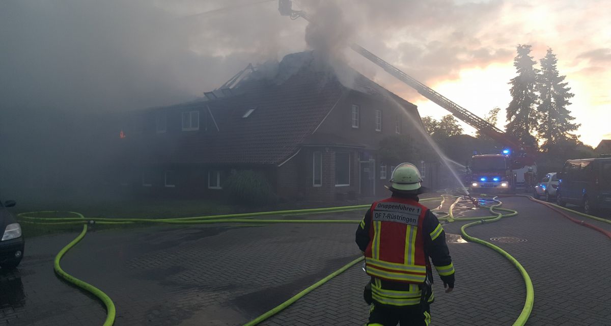 Auf dem Foto ist ein Haus zu sehen. Das Haus ist mit starkem Rauch umhüllt. Es brennt das Dach. Die Feuerwehr löscht es aktuell. Vorne ist ein Mann ist Uniform zu sehen.