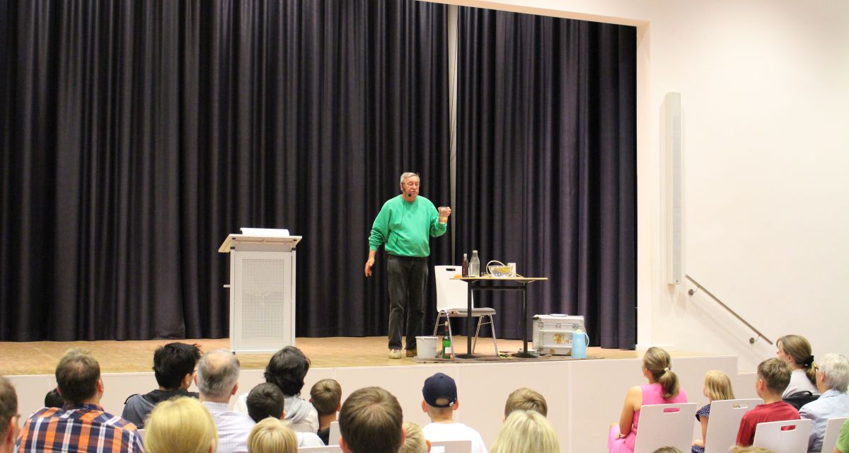 Christoph Biemann steht auf einer Bühne in der Aula und führt Experimente durch