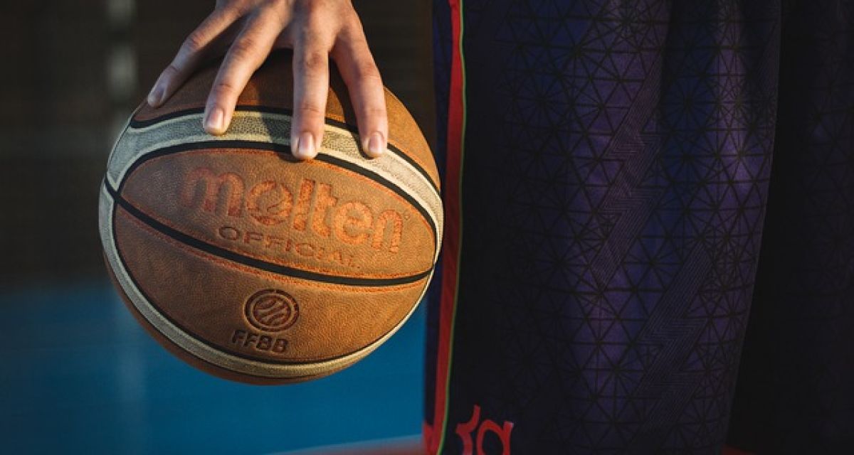 Zu sehen ist die Hand eines Mannes, die einen Basketball hält. Außerdem ist zu sehen, dass der Mann eine kurze Sporthose trägt und in einer Sporthalle steht.