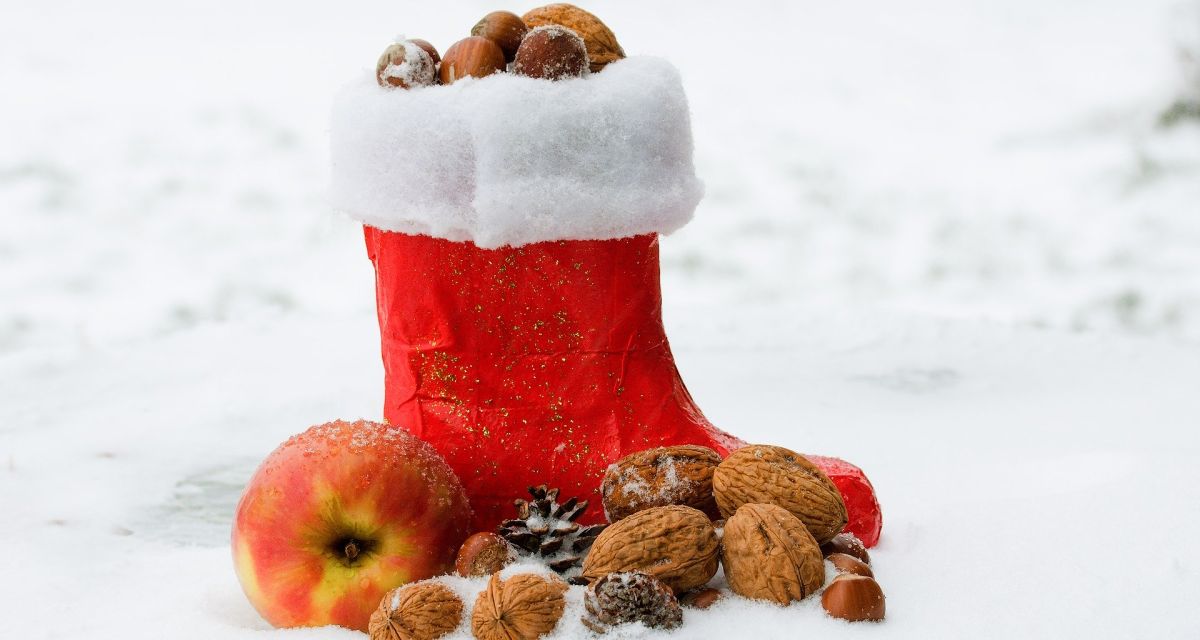Zu sehen ist ein roter Nikolaus-Stiefel mit weißem Pelzsaum. Gefüllt ist dieser Stiefel mit Nüssen. Um den Stiefel herum liegen weitere Nüsse und ein Apfel im Schnee.