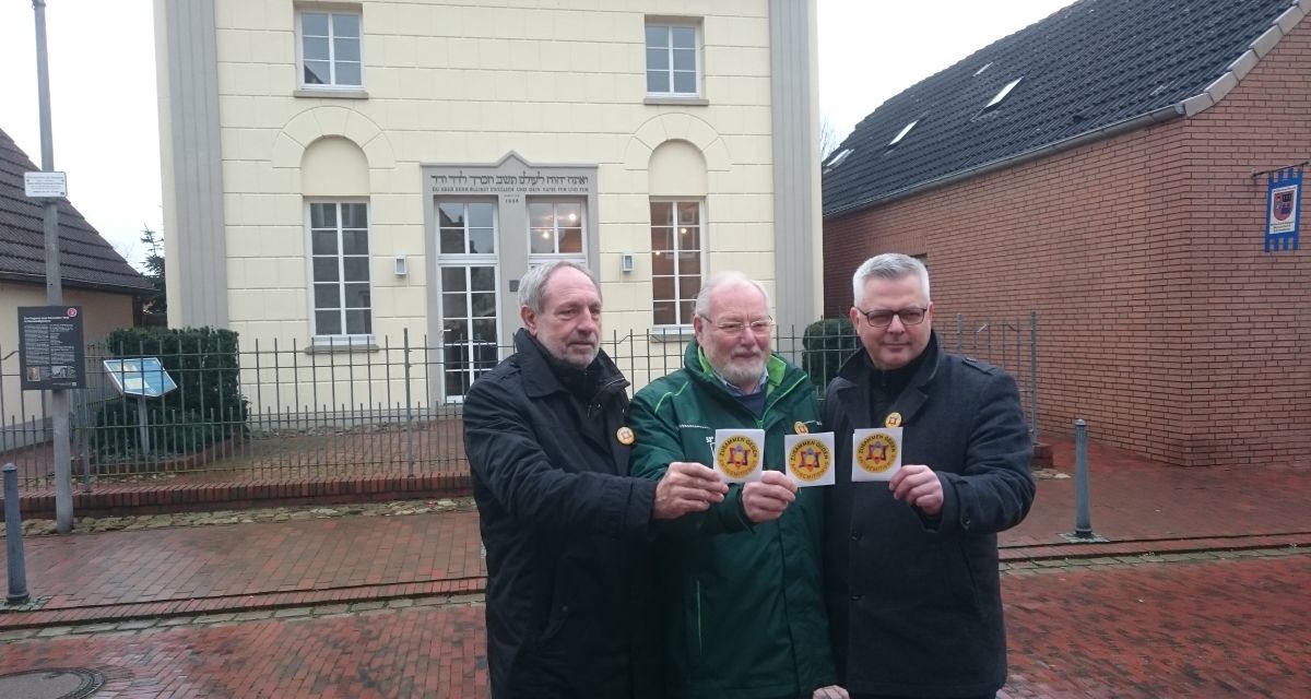 Stephan Eiklenborg hält zusammen mit zwei weiteren Personen einen Sticker in die Kamera. Im hintergrund steht die Synagoge in Neustadtgödens