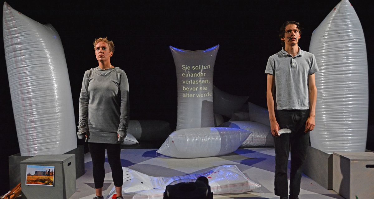Eine Frau und ein Mann stehen auf einer Bühne. Sie tragen graue Kleidung und schauen nach vorne.