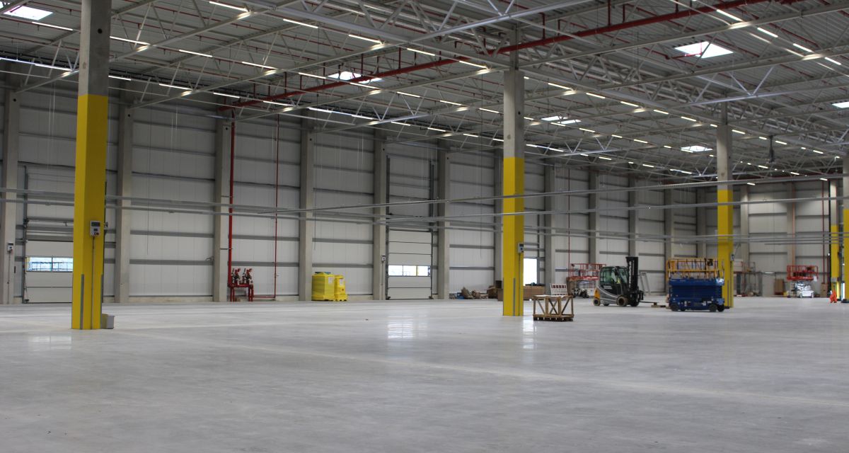 Das leere neue Logistikzentrum von Amazon Logistics zum Zeitpunkt des Richtfests. Es ist eine große leere Halle mit glänzendem Boden und grau/gelben Trägern. 