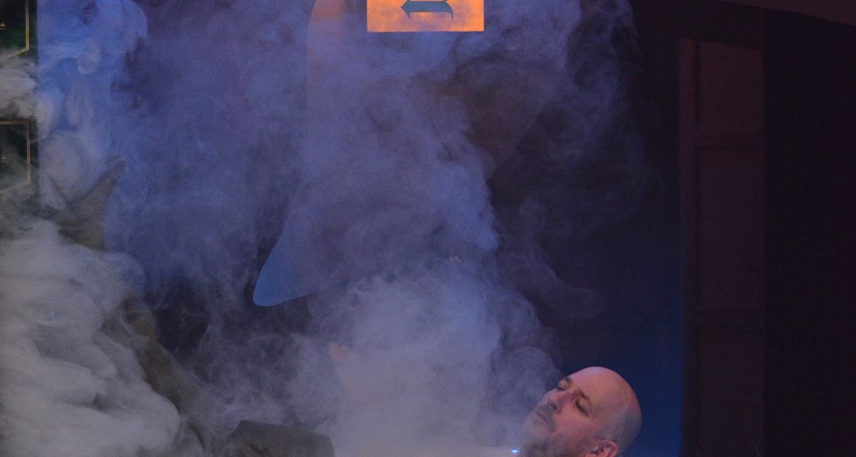 Ein Mann liegt auf einer Bühne in einer Badewanne. Um ihn herum ist Rauch, über ihm hängt ein Schild