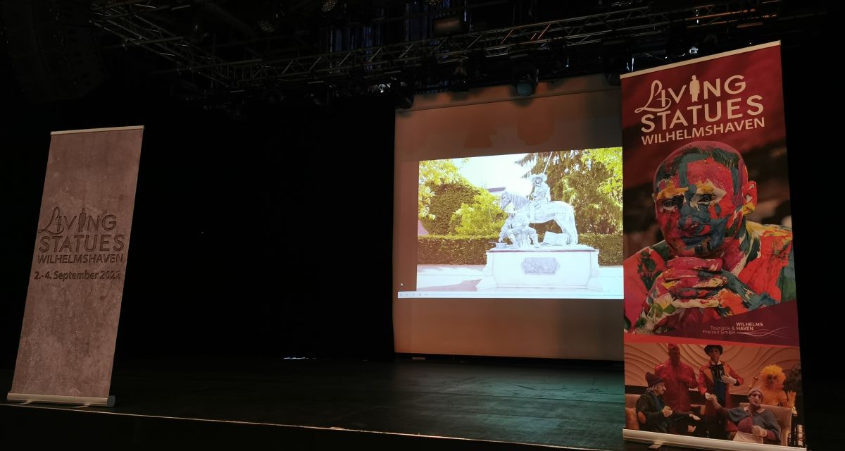 Auf dem Foto ist eine dunkle Bühne zu sehen. Im Hintergrund ist eine Reiterstatue an eine Wand angestrahlt, rechts und links stehen Aufsteller für das Living-Statues-Festival in Wilhelmshaven