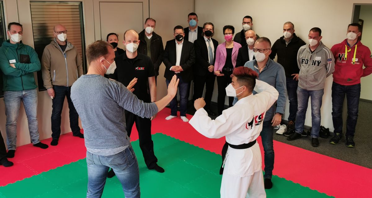 Im Rahmen des Pilotprojekts zur Selbstbehauptung bei Feuerwehrkräften demonstrieren zwei Männer Handgriffe zur Selbstverteidigung. Sie stehen auf einer grünen Matte, ringsum stehen Zuschauer*innen.