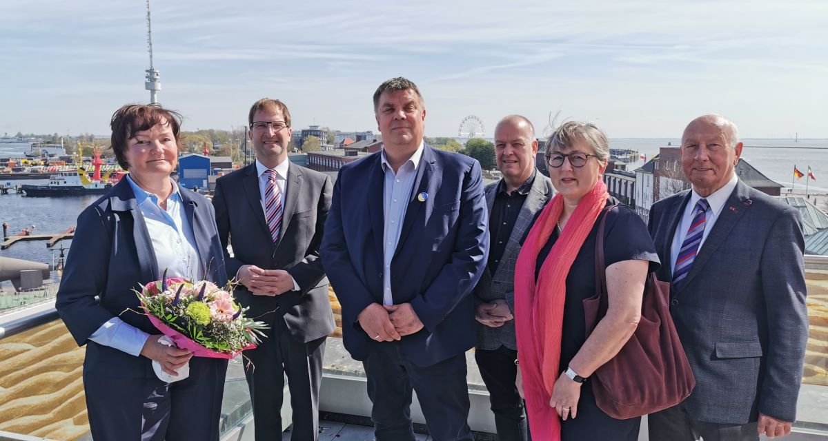 Zu sehen ist die scheidende Leiterin des Eigenbetriebs Gebäude und Grundstücke der Stadt Wilhelmshaven, Simone Groh, bei ihrer Verabschiedung. Neben ihr stehen Oberbürgermeister Carsten Feist, Stadtbaurat Niksa Marusic sowie weitere Vertreter:innen der Stadtpolitik. Im Hintergrund ist der Hafen zu sehen.