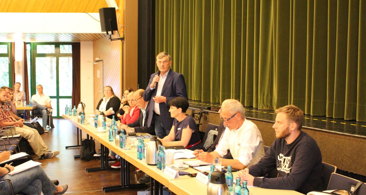 Vertreter*innen von Stadt Schortens und Landkreis Friesland sitzen an vor einem Publikum. Bürgermeister Gerhard Böhling steht und hält ein Mikrofon in der Hand