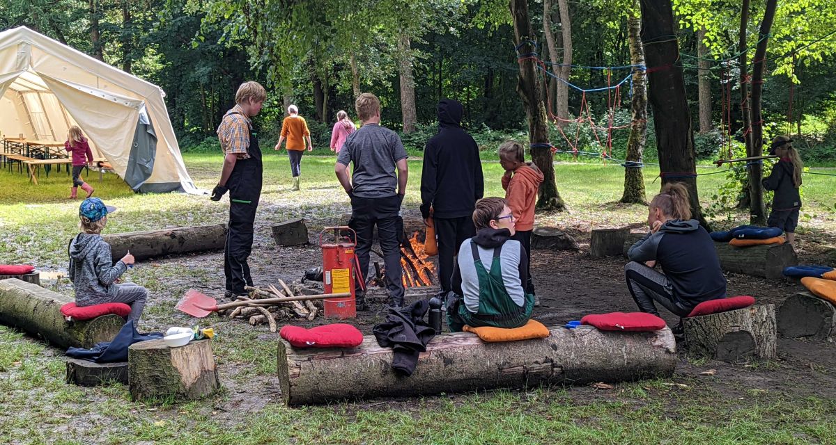 Kinder sitzen beim NEO-Camp Jever auf Holzstämmen um ein Feuer, einige Erwachsene stehen dabei. 