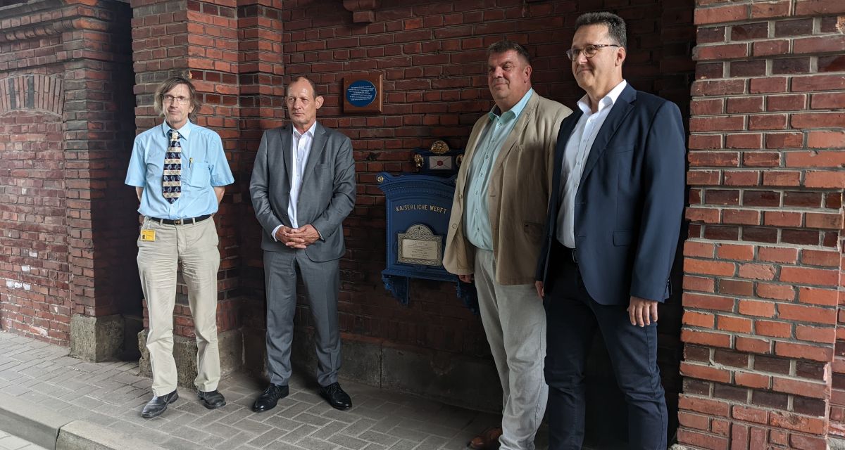 Der historische Briefkasten am Tor 1 des Wilhelmshavener Marinearsenals wird nach seiner Sanierung enthüllt. Zu sehen sind unter anderem Oberbürgermeister Carsten Feist, der leitende Direktor des Marinearsenals Rainer Sacher und ein Vertreter der Deutschen Post. Sie stehen vor dem Briefkasten, über dem außerdem eine blaue Erklärungsplakette hängt.