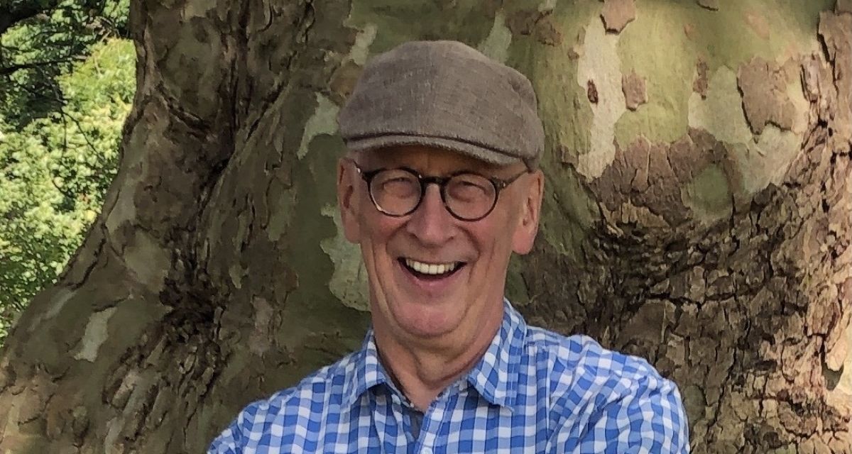 Pastor Frank Morgenstern trägt eine Mütze, steht vor einem Baum und lacht herzlich in die Kamera.