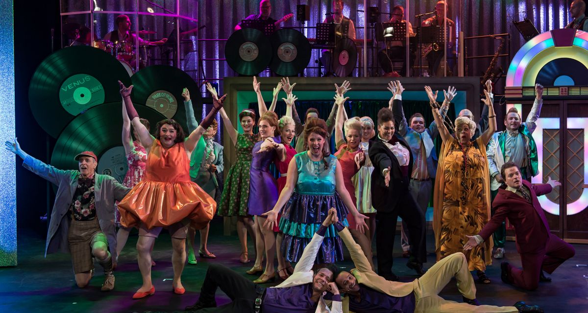 Das Ensemble des Musicals Hairspray steht in Pose auf der Bühne. Alle tragen bunte Kostüme