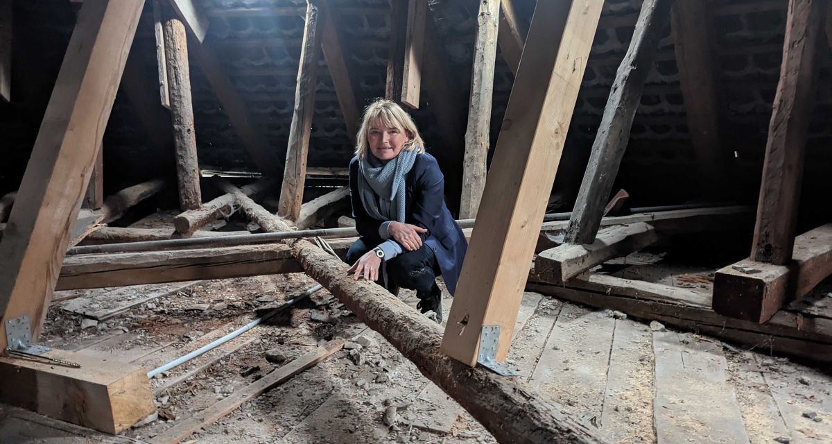 Die Leiterin des Schlossmuseums Jever, Dr. Antje Sander, hockt auf dem Dachboden des Eulenturms des Schlosses. Der sie umgebende Dachstuhl besteht aus alten und neuen Holzstreben, die mit Metallklammern verbunden sind. Durch Dachluken im Hintergrund fällt Licht.
