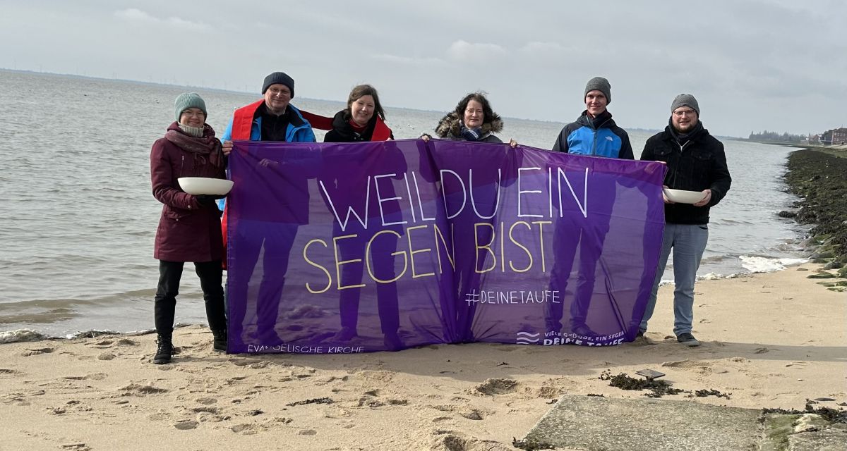 Pastor*innen stehen am Südstrand und halten ein Banner hoch, auf dem steht