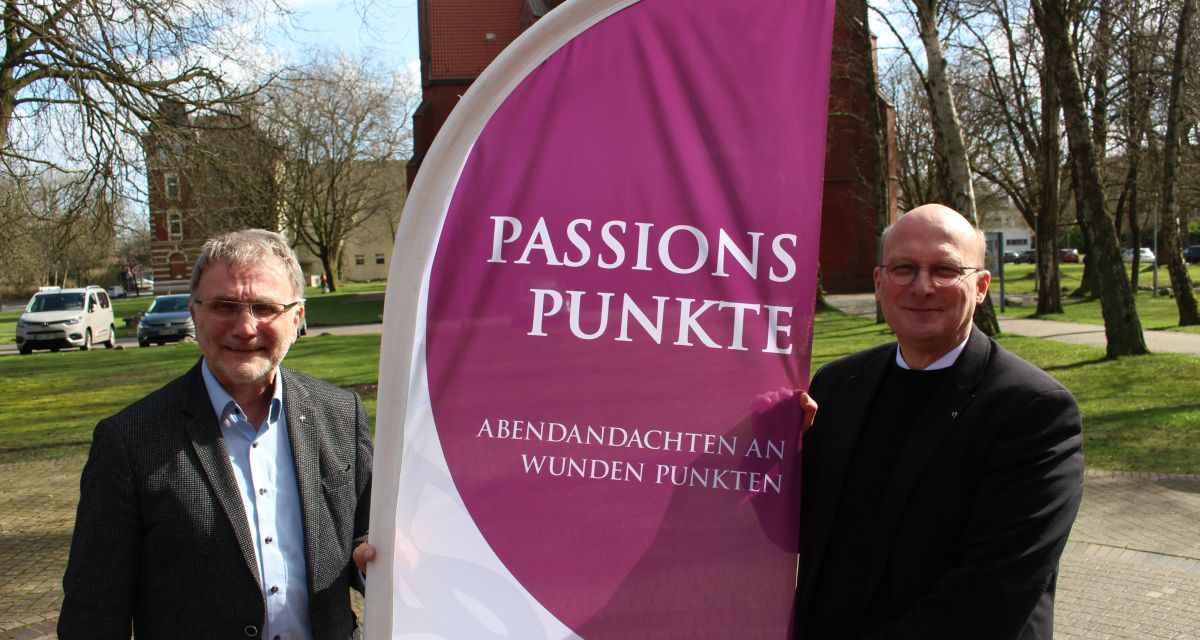 Die Passionspunkte 2023 werden vorgestellt. auf dem Kirchplatz der Christus- und Garnisonkiche in Wilhelmshaven zu sehen sind Gemeindekirchenrat Rüdiger Schaarschmidt und Pastor Bernhard Busemann. Sie halten eine Flagge mit dem Logo der Passionspunkte und dem Slogan 