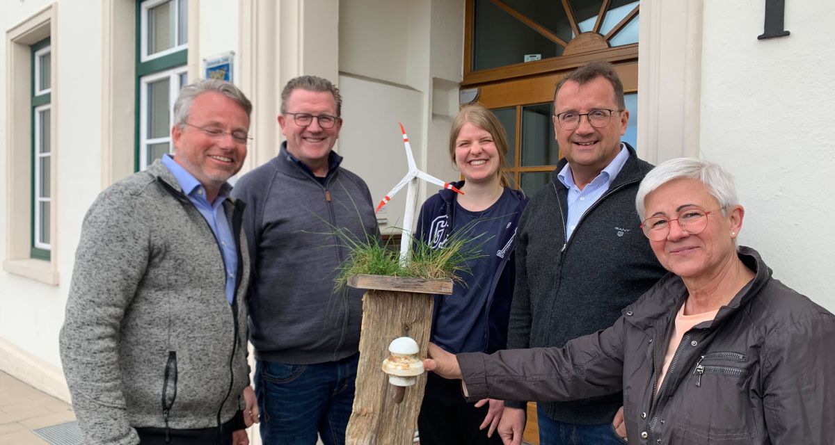 Vertreter*innen der Klimagruppe Friesische Wehde und der Gemeinde Zetel stehen vor dem Rathaus um einen Zaunpfahl, auf dem eine Modell-Windkraftanlage befestig ist