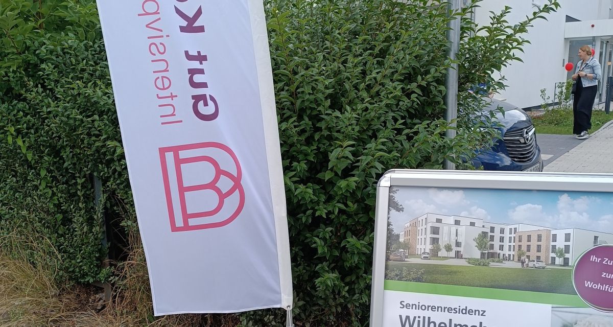 Die neue Intensivpflege-Wohngruppe des Betreibers Gut Köttenich Intensivpflege in Wilhelmshaven wird an der Marktstraße vorgestellt. Zu sehen ist eine Beachflag des Betreibers und ein Aufsteller, der über die Einrichtung informiert. Im Hintergrund ist der Eingang des Gebäudes zu erkennen.