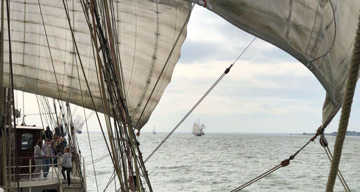 Zu sehen ist ein Ausschnitt eines Segelschiffes auf dem Meer, sowie weitere Schiffe, die im Hintergrund auf dem Meer fahren. 