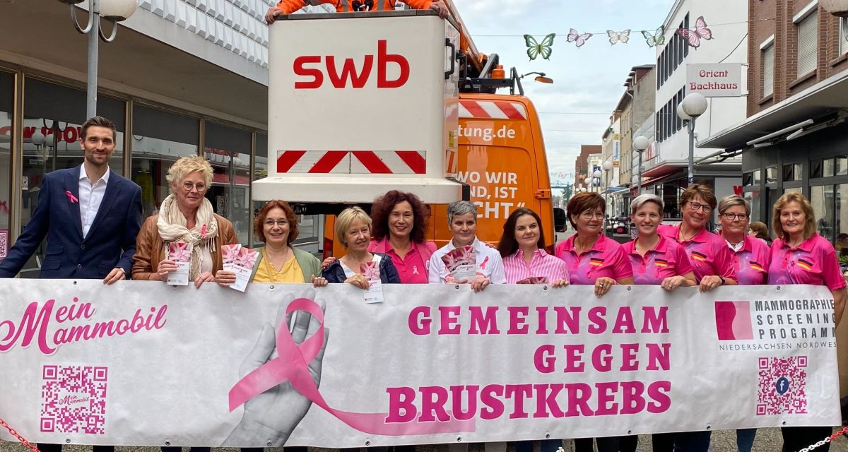 Veranstalter*innen des Brustkrebsfrüherkennungsmonats Pinktober stehen mit einem Transparent vor dem Kaufhaus Leffers in Wilhelmshaven. Im Hintergrund ist die aufgehängte Kette aus pinken Büstenhaltern zu sehen, auf dem Transparent steht in pinker Schrift 
