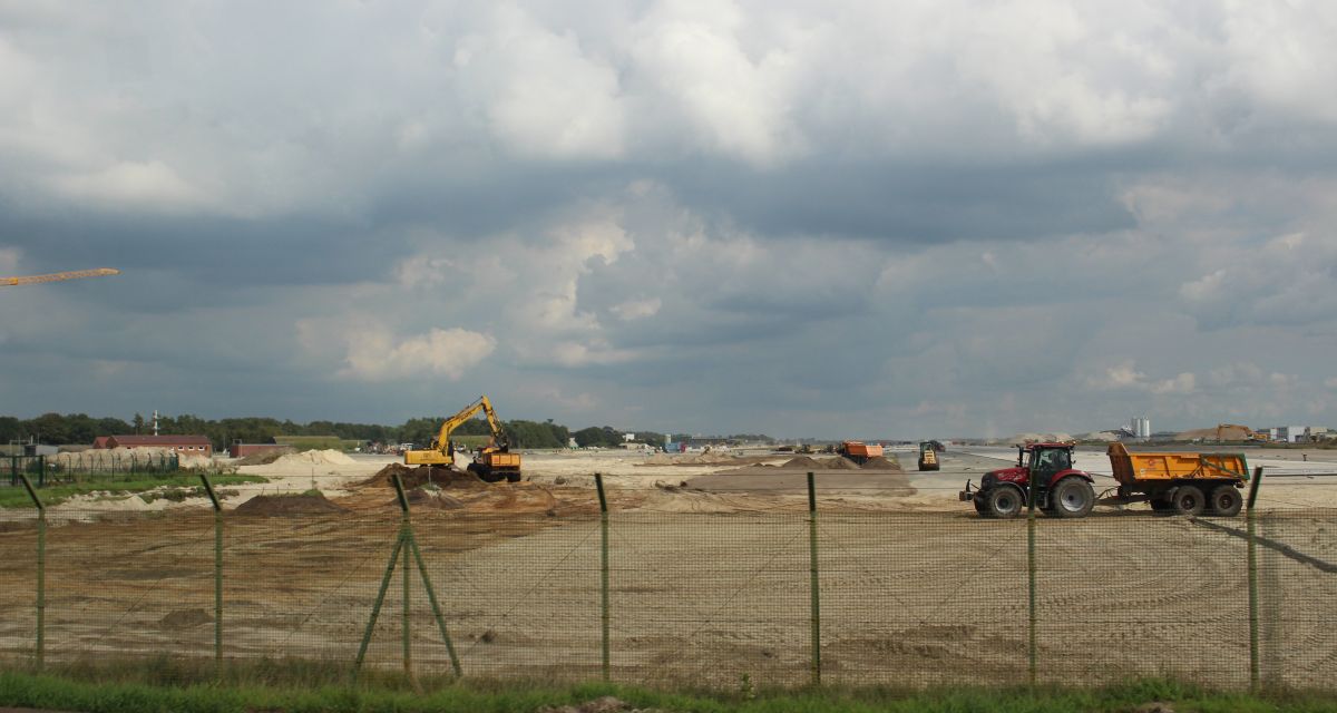 Zu sehen ist eine große freie Fläche, auf der Bagger und andere Baugeräte verschiedene Arbeiten verrichten. 