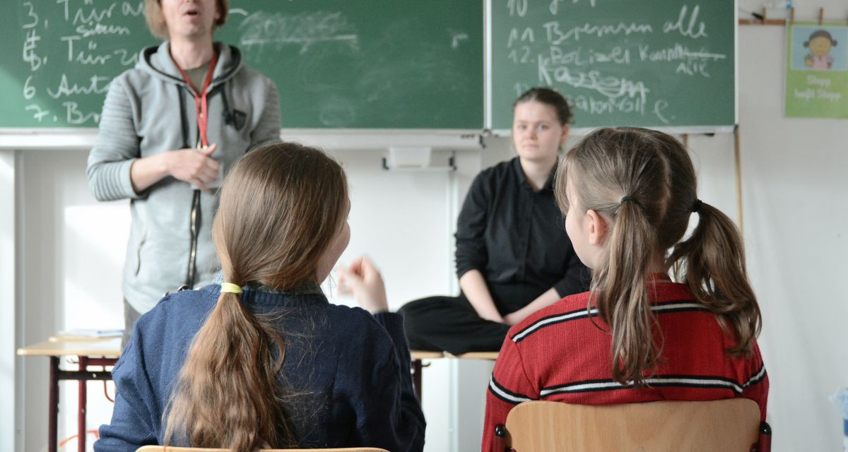 Zwei Kinder sitzen auf Stühlen in einem Klassenraum. Vor ihnen stehen zwei Personen vor einer Tafel.