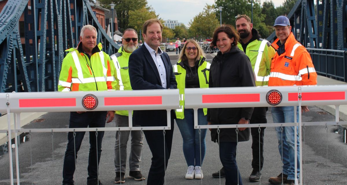 Die Wiedereröffnung der frisch restaurierten Deichbrücke in Wilhelmshaven für den gesamten Verkehr. Hinter der noch geschlossenen Schranke stehen Stadtbaurat Niksa Marusic und TBW-Betriebsleiterin Maike Schun mit Mitarbeitenden der Technischen Betriebe Wilhelmshaven in gelben Schutzwesten.