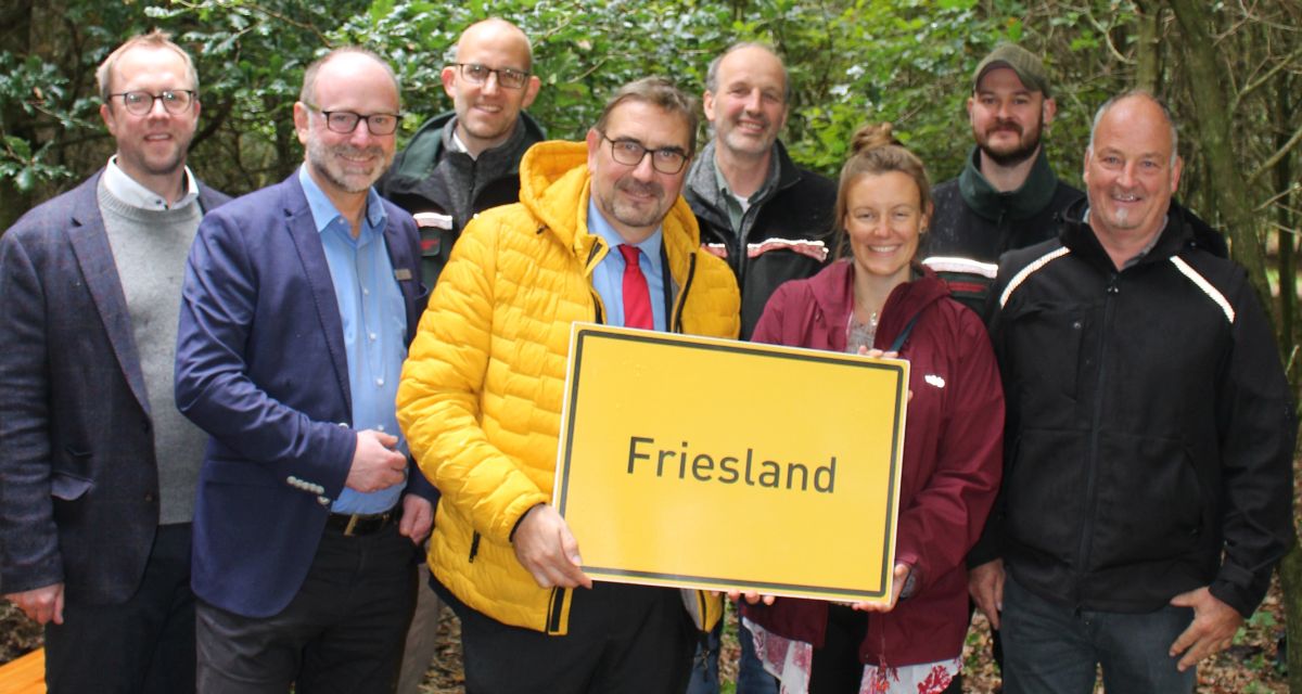 Im Bild zu sehen sind Markus Neumann, Klaus Blum, Uwe Mestermacher, Sven Ambrosy, Tido Bent, Dr. Ilka Strubelt, Fiddy Winkelhake und Andreas Schmidt vor jungen Bäumen im Wald bei Varel. Sie halten ein Schild mit der Aufschrift