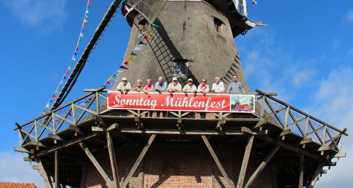 Zu sehen ist die Schlachtmühle in Jever vor blauem Himmel. Auf der Galerie stehen einige Mitglieder des Abeitskreises Schlachtmühle aus dem Jeverländischen Altertums- und Heimatverein.
