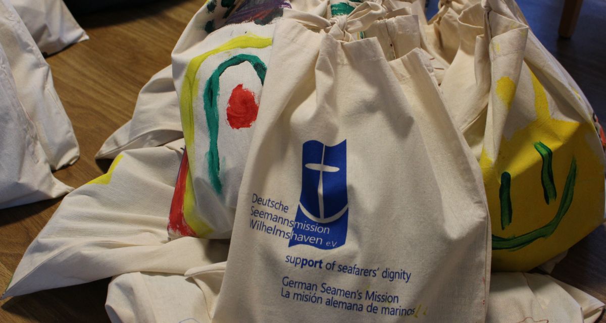 Auf dem Bild sind Jute-Beutel zu sehen, die bunt bemalt sind und mit dem Logo und Schriftzug der Deutschen Seemannsmission Wilhelmshaven geprägt sind.