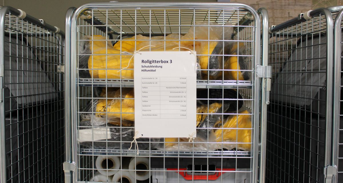 Zu sehen ist einer der sechs Notfallcontainer und eine Aufführung seines Inhalts. Außerdem lassen sich einige Gegenstände im Container erkennen: Gummistiefel; aufgerollte Folie; eine Kiste mit Handschuhen und Warnwesten.