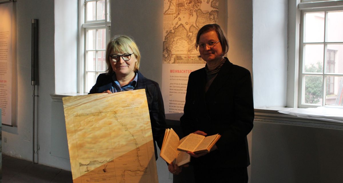 Auf dem Foto sind die Organisatiorinnen der Ausstellung zur Februarflut 1825 zu sehen: Dr. Antje Sander (links, Leiterin des Schlossmuseums Jever) und Christiane Baier (rechts, Leiterin der Bibliothek und des Archivs des Schlossmuseums). Antje Sander hält eine historische Karte, auf der die friesische Küste zu sehen ist, auf der die Überschwemmungen der Flut eingetragen sind. Christiane Baier hält zwei Bücher mit Aufzeichnungen der Geschehnisse von 1825.
