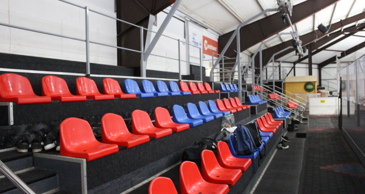 Zu sehen sind Zuschauerränge in der Eishalle Sande. Rote und blaue Plastikschalensitze sind fest auf Stufen montiert. Rechts ist die Trennwand zur Eisfläche zu sehen, im Hintergrund ist das Unternehmenslogo der Jever Brauerei zu erkennen.