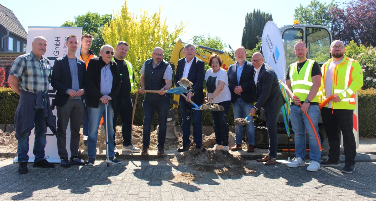 Spatenstich für den Glasfaserausbau im Schortenser Ortsteil Grafschaft. Auf dem Gehweg in der Moorsumstraße stehen Vertreter*innen der Schortenser Politik (unter anderem Bürgermeister Gerhard Böhling, stellvertretender Bürgermeister Axel Homfeldt und Ratsvorsitzende Heide Bastrop), der Deutschen Glasfaser (unter anderem Senior Manager Kommunale Kooperation Michael Sieve) und der ausführenden Firma Libra GmbH. Mit Spaten wird symbolisch Sand auf die Straße geworfen, im Hintergrund steht ein Bagger.