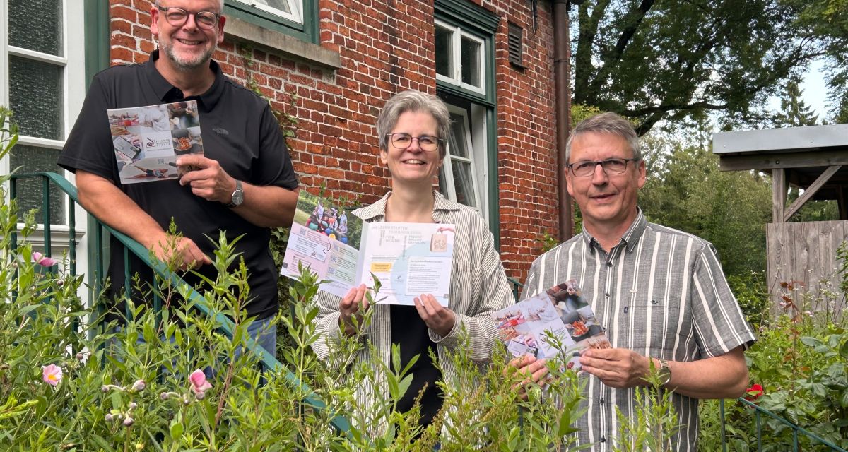 Zu sehen sind drei Personen vor einem Hauseingang hinter Blumen auf einer Treppe: Kreispfarrer Christian Scheuer, EFB-Leiterin Claudia Lehniort und Pastor Thorsten Harland aus Jever