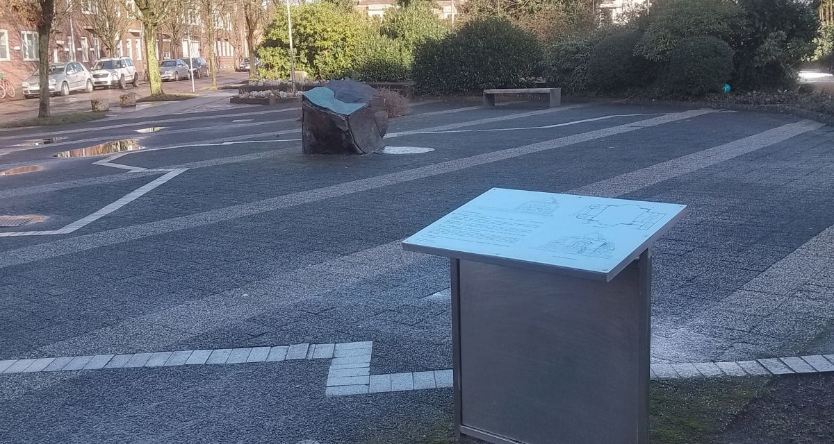 Der Synagogenplatz in Wilhelmshaven. Im Vordergrund ist eine Informationstafel zu erkennen, im Hintergrund ein Gedenkstein. Auf dem Boden sind die Umrisse der in der Pogromnacht 1938 zerstörten Synagoge in weißen Pflastersteinen nachempfunden.