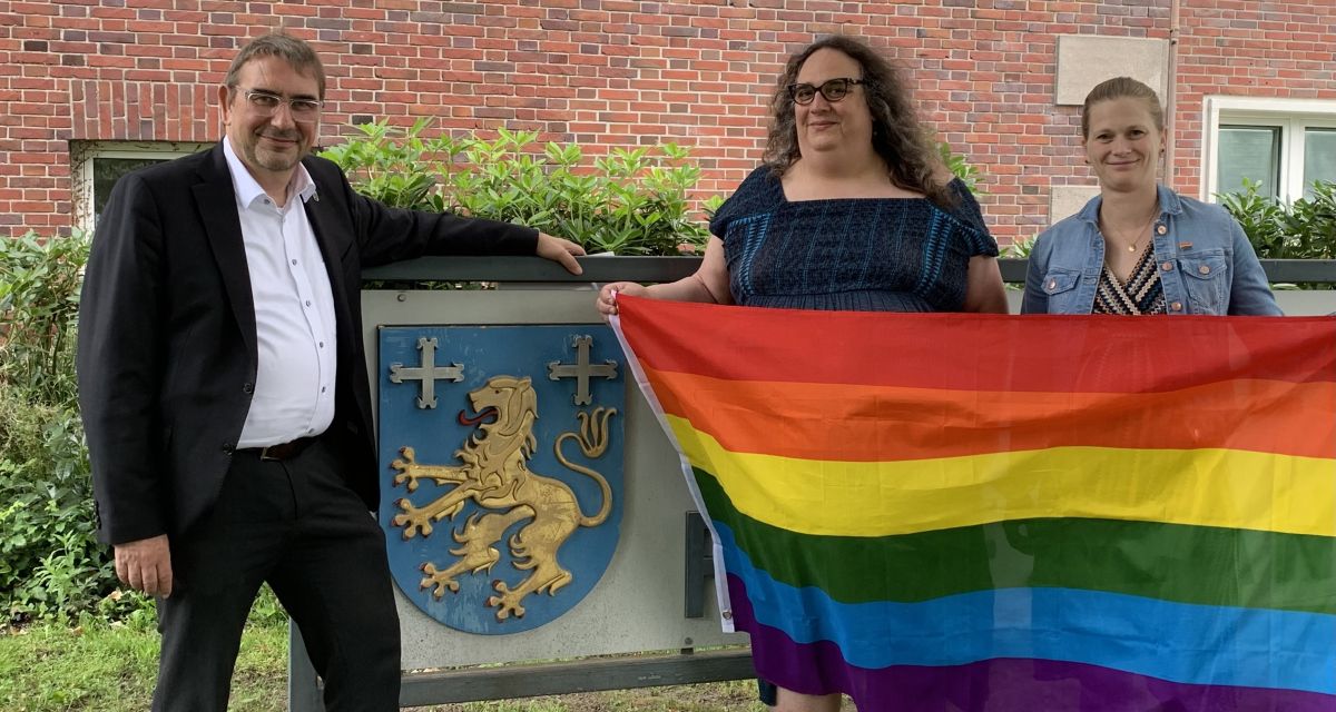 Die Queere Beratungsstelle im Landkreis Friesland wird wieder besetzt. Durch einen Trägerwechsel war die Stelle einige Zeit vakant. Auf dem Bild zu sehen sind (von links): Landrat Sven Ambrosy, Freyja Pe* von Rüden (Queere Beratungsstelle Friesland) und Gleichstellungsbeauftragte Ann-Kathrin Cramer. Sie stehen vor dem Schild des Landkreises Friesland, zu sehen ist der friesische Löwe auf dem Wappen. Außerdem halten sie eine große Regenbogenflagge, als Symbol für die queere Community. 