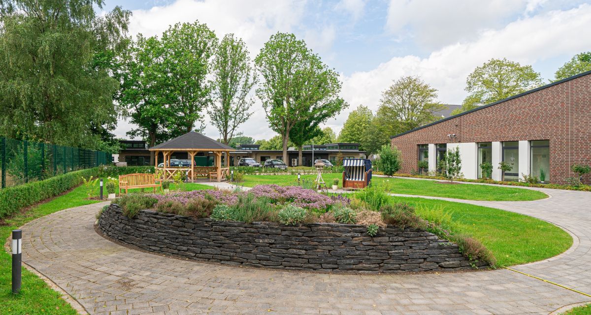 Zu sehen ist der Garten des Hospizes am Wattenmeer in Varel. Gepflasterte Wege ziehen sich durch Rasenflächen, darin ein gemauertes Beet, ein Strandkorb, ein Vogelhäuschen, eine Bank und eine Pagode.