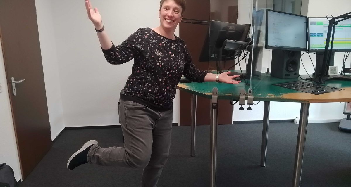 Die Kabarettistin und Slam-Poetin Annika Blanke im Hörfunkstudio C von Radio Jade in Wilhelmshaven. Sie steht auf einem Fußhocker auf einem Bein und lächelt in die Kamera.