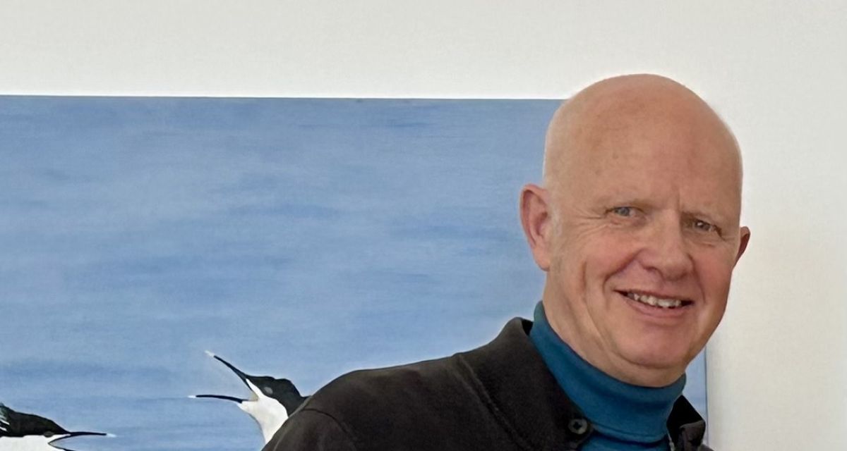 Zu sehen ist ein haarloser älterer Herr vor einem Bild,d as einen Vogel zeigt.