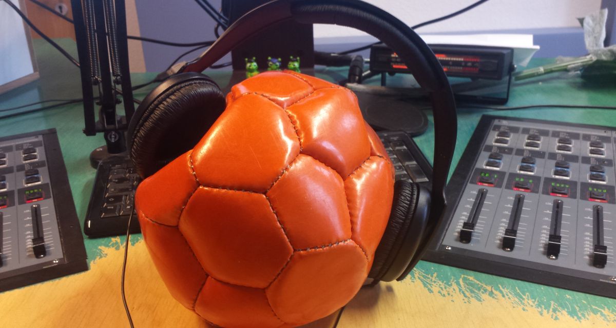 Ein orangener Ball, dem ein Studiokopfhörer aufgesetzt wurde. Im Hintergrund die Pulte des Studios B bei Radio Jade.