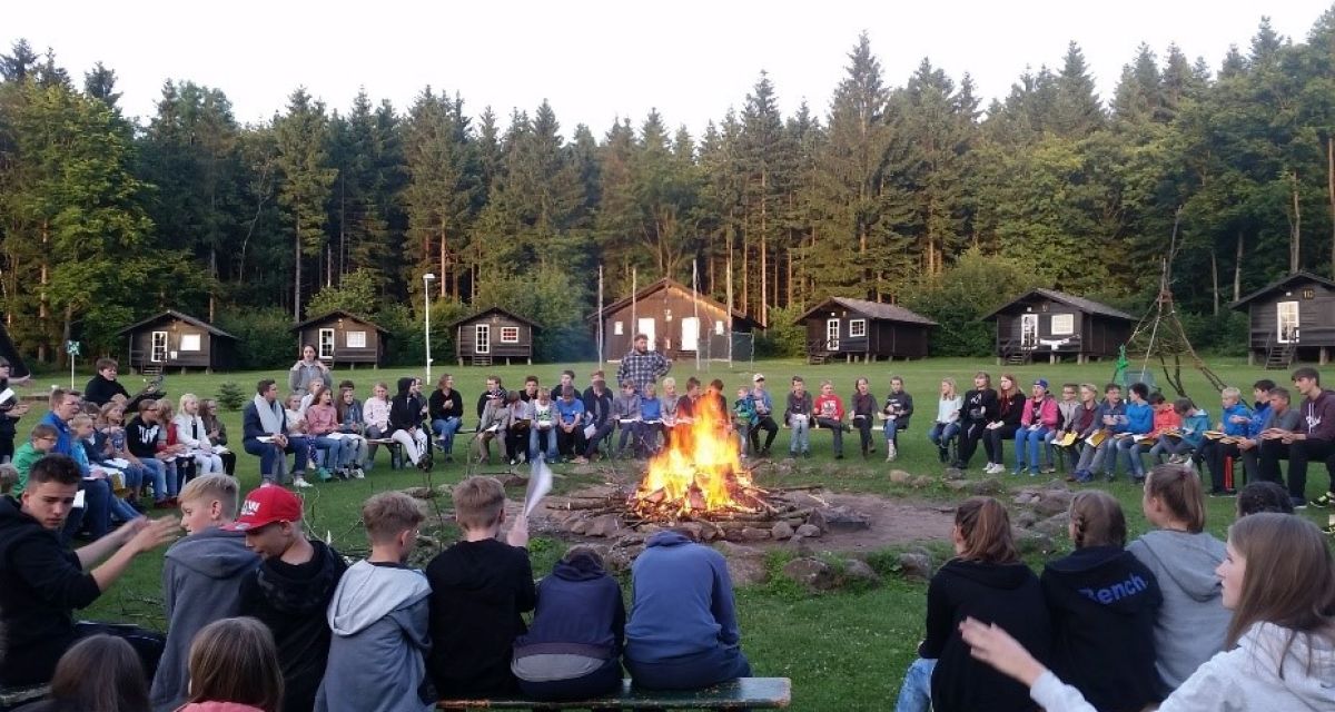 Zu sehen ist ist eine große Gruppe vorwiegend jugendlicher Menschen am lagerfeuer in einem Hüttendorf