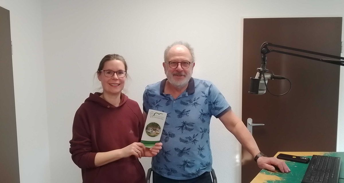Katharina Glaum und Wolfgang Schuy vom Naturschutzhof Wittmunder Wald zu Besuch im Studio von Radio Jade. Die beiden haben über Kochkurse im Naturschutzhof im Jahr 2026 informiert. Sie stehen am Sendepult von Studio B, Glaum hält eine Informationsbroschüre in der Hand. Auf der rechten Seite ist ein Mikrofon zu sehen.