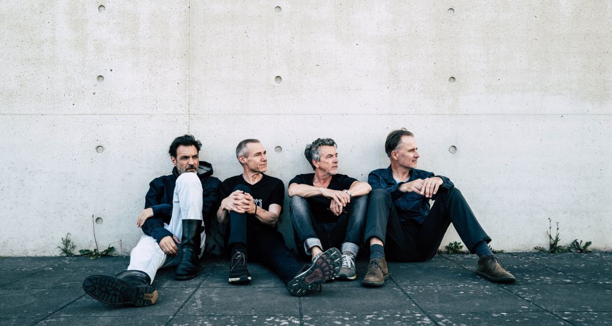 Zu sehen ist die Band vor einer Betonwand sitzend ohne Instrumente