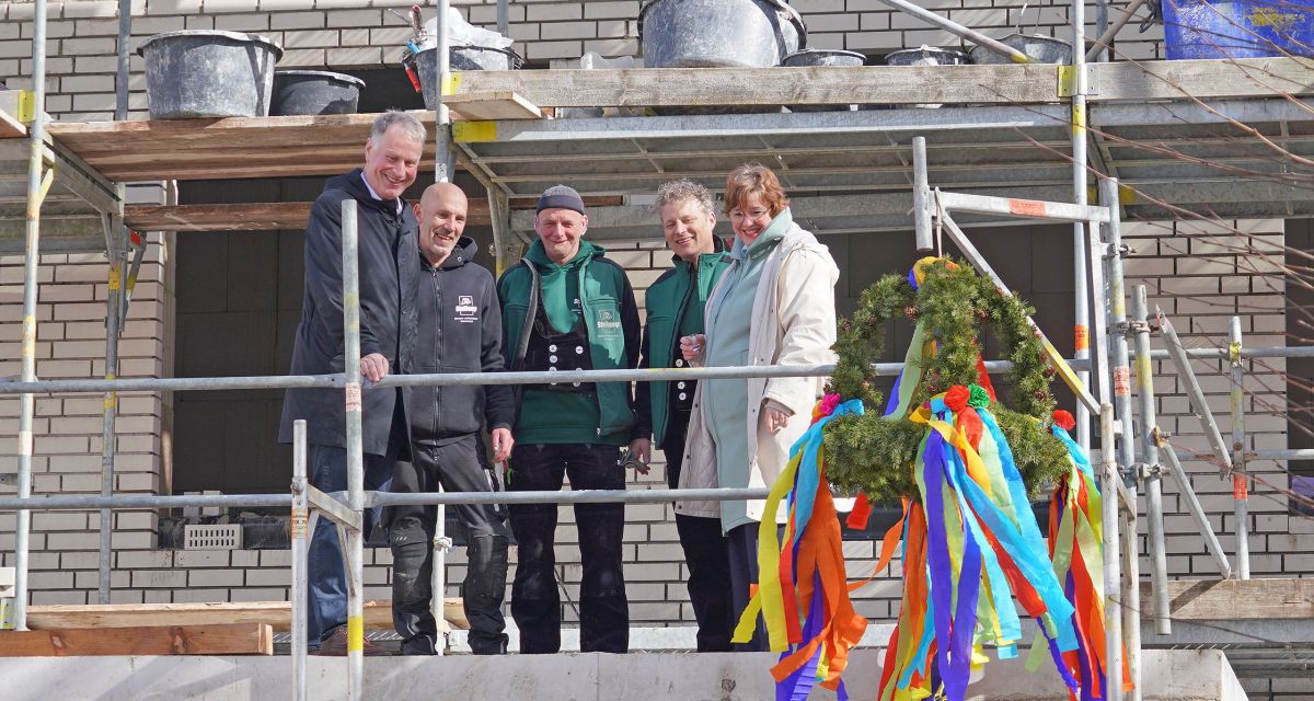 Am Neubau der Grundschule an der Hafenstraße in Varel wird Richtfest gefeiert. Das Bild zeigt auf dem Gerüst Bürgermeister Gerd-Christian Wagner und die kommissarische Schulleiterin Miriam Engels-Isigwe mit Vertretern des Unternehmens Stallkamp. Im Vordergrund ist der bunte Richtkranz aufgehängt, auf dem Gerüst stehen außerdem noch dunkelgraue Speisfässer.