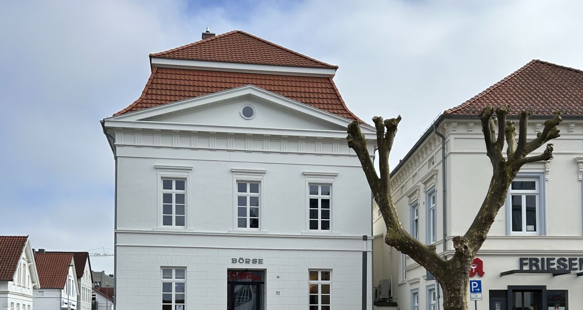 Zu sehen ist ein weißes zweistöckiges Gründerzeitgebäude an einer Straßenecke in Varel, die Börse der Ideen. Davor steht eine runde Sitzbank