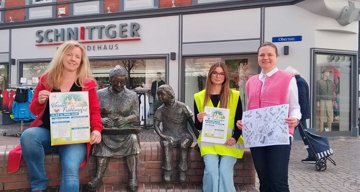 In Varel wird das Programm des Frühlingsfestes 2026 vorgestellt. Am Denkmal der Krabbenpulerin in der Obernstraße stehen (von links nach rechts) Meike Knop, die Geschäftsführerin des Stadtmarketings, Noémie Fernandez vom Jugendzentrum Weberei und Marktmeisterin Yvonne Uhr. Sie halten Plakete des Fests und einen Lageplan in den Händen.