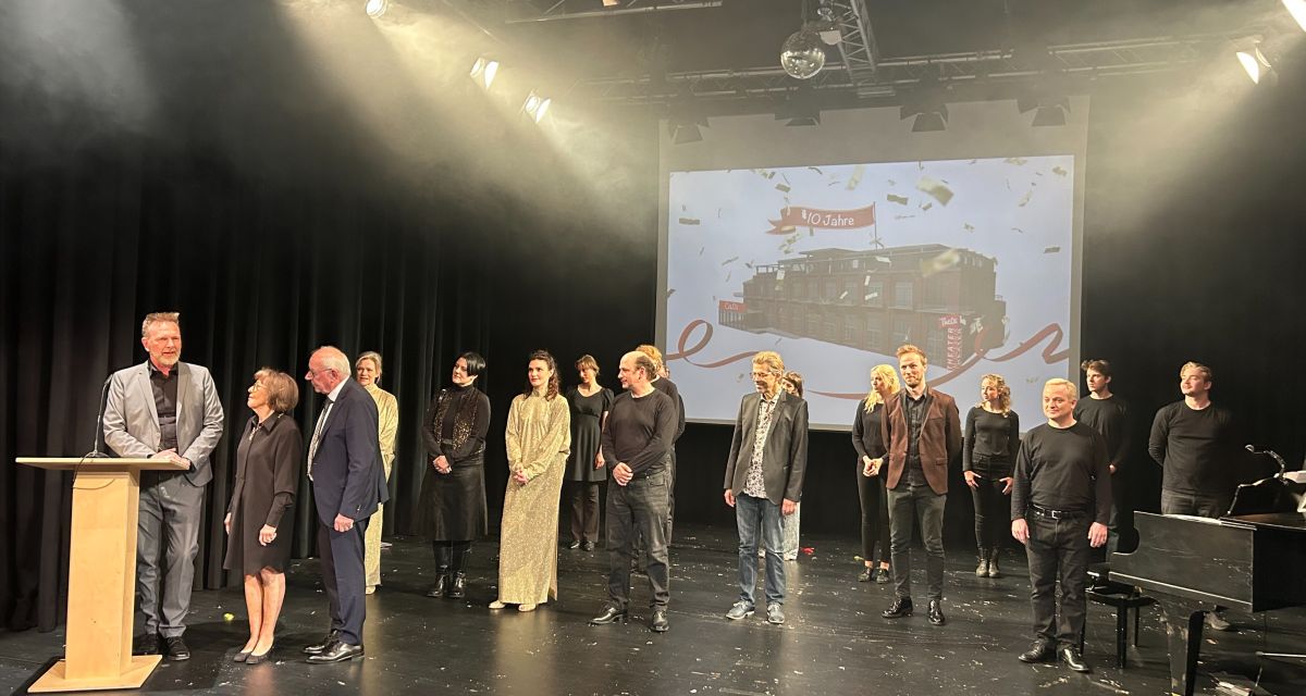 Zu sehen ist die Bühne des Theater am Oceanis (TheOs) in Wilhelmshaven. auf ihr stehen anlässlich des Festaktes zum zehnjährigen Bestehen Mitglieder des Ensembles und Mitarbeitende der Landesbühne Niedersachsen Nord, Intendant Olaf Strieb und Mäzenin Angelika Reichelt. Auf einer Leinwand im Hintergrund ist ein Bild des TheOs mit dem Banner 