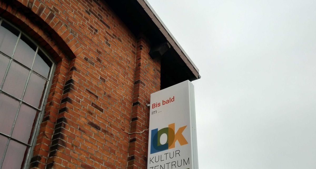 Zu sehen ist eine Ecke der Stirnseite des LOK Kulturzentrums in Jever. An der roten Klinkermauer hängt ein Schild mit der Aufschrift