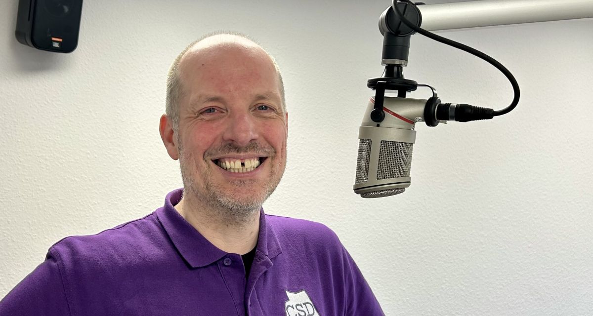 Zusehen ist im Radiostudio ein lächelnder mittelalter Mann im Lila CSD-Poloshirt