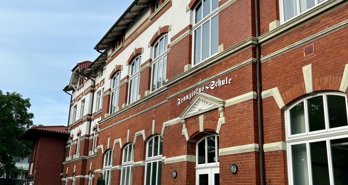 Zu sehen ist die Fassade eines Schulgebäudes aus der Kaiserzeit iin der Schrägansicht. Über dem Eingang ist der Schriftzug